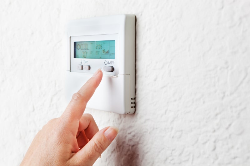 A hand reaching toward a button on a thermostat, hung on a white wall